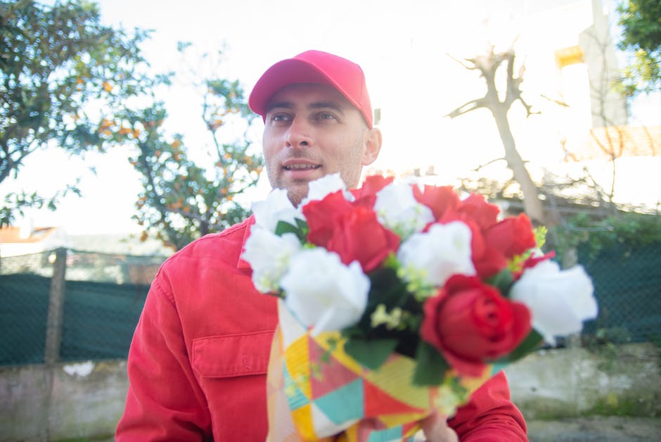 A man dressed in a red jacket and matching red cap is holding a vibrant mixed bouquet of fresh flowers, including red and white roses, wrapped in colorful patterned paper. He is outdoors, with a backg