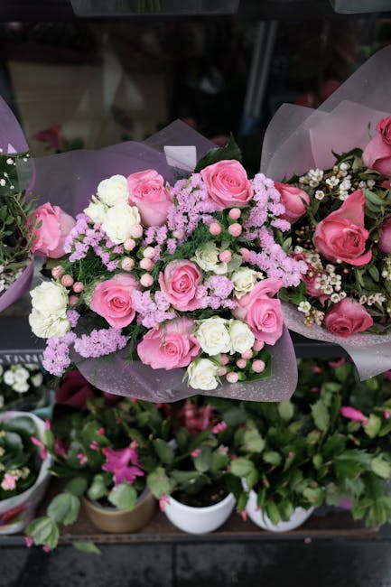 A floral arrangement featuring a bouquet of fresh pink roses, white carnations, and small pink and white filler flowers, all set within lavender translucent wrapping paper and tied with a delicate pin