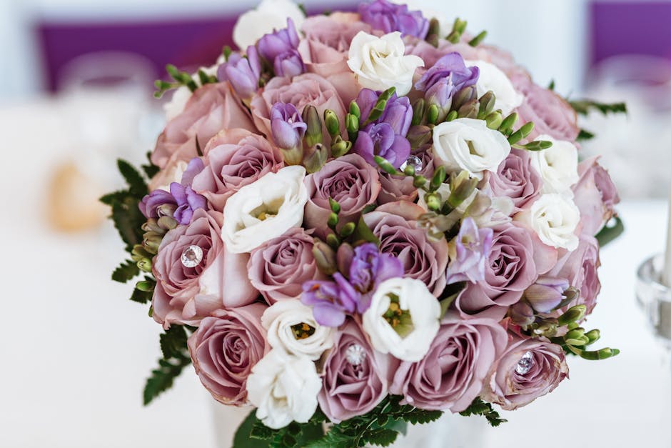 A close-up of a floral bouquet featuring soft pink roses, white lisianthus, and purple freesias, accented with small green buds and dark green foliage. The arrangement is densely packed, showcasing th