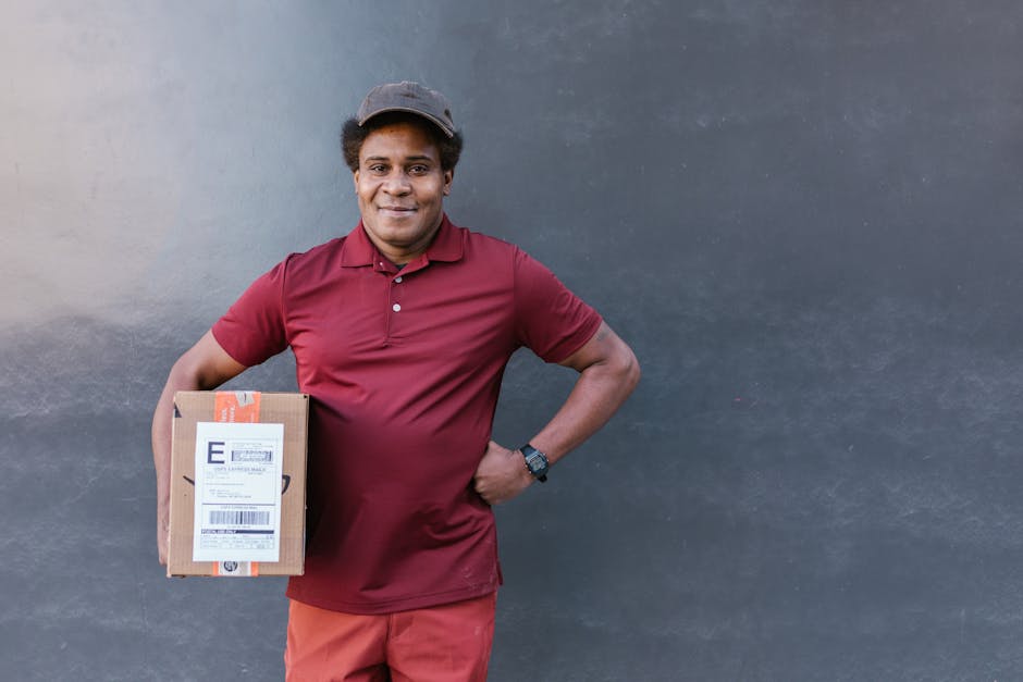 A friendly delivery person, with medium skin tone and curly hair, stands outdoors against a plain, two-tone background wall. He is wearing a maroon polo shirt, matching maroon pants, and a dark-colore