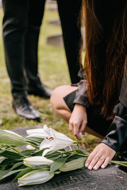 A florist, dressed in dark clothing, is kneeling on the ground placing a white lily bouquet wrapped in clear paper and tied with a ribbon, which is arranged with lush green leaves. The bouquet rests o
