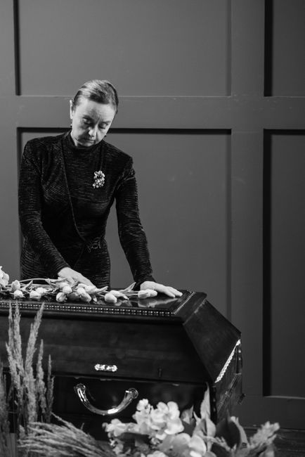 A woman dressed in black, with her hair pulled back, is arranging a floral tribute on a polished wooden casket during a funeral ceremony. She is carefully placing white roses and greenery, which are p